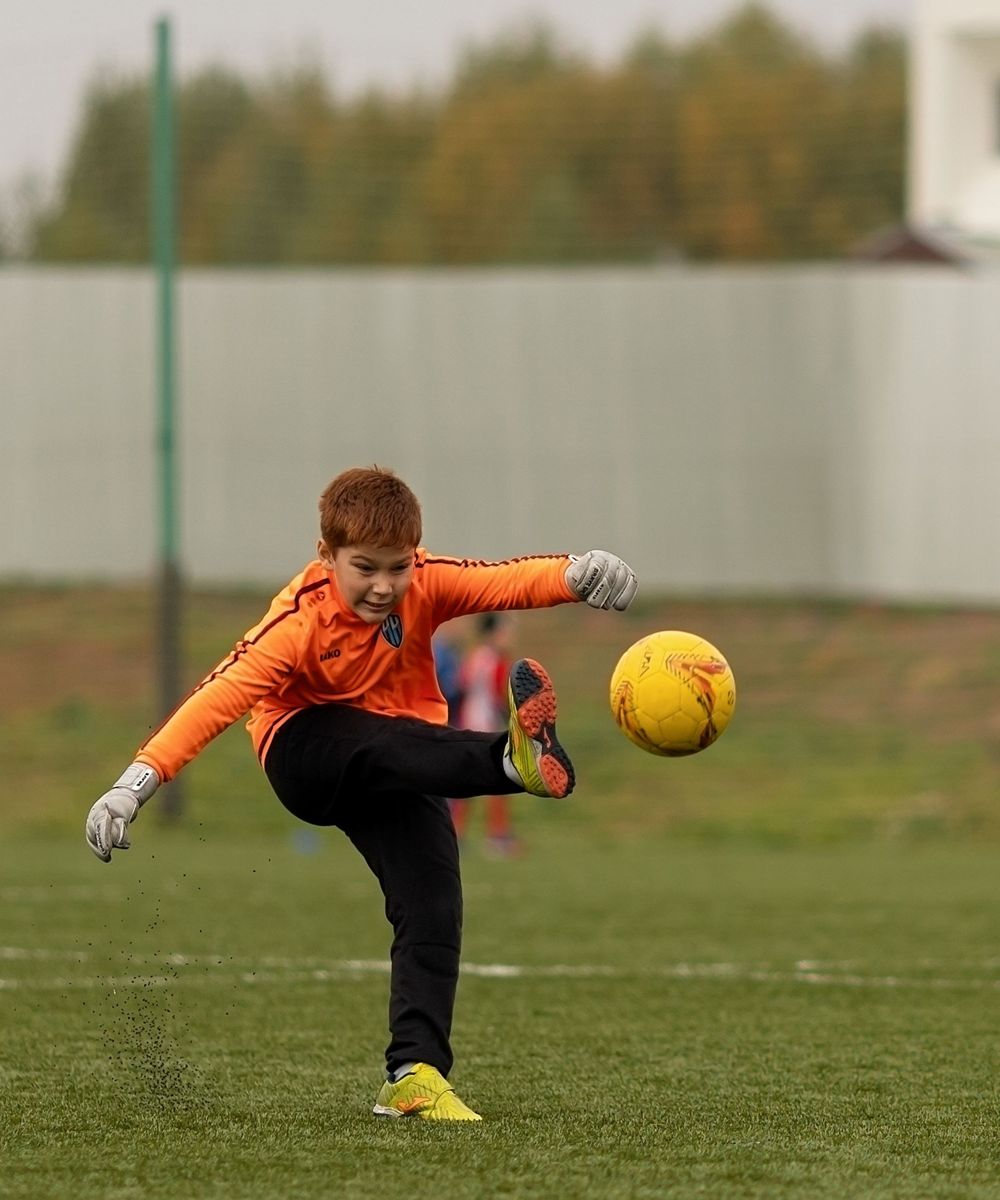 Un jeune gardien en maillot orange dégage le ballon sur un terrain de club, en automne